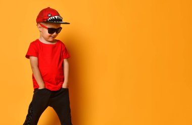 Kırmızı tişörtlü, siyah pantolonlu, beyaz spor ayakkabılı ve sarı arka planda bedava fotokopi alanında poz veren komik şapkalı çocuk.