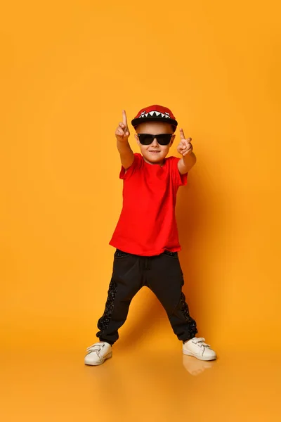 Młody chłopak w czerwonej koszulce i ciemnych spodniach, białych trampkach i śmiesznej czapce, pozujący na wolnej przestrzeni na żółtym tle. — Zdjęcie stockowe