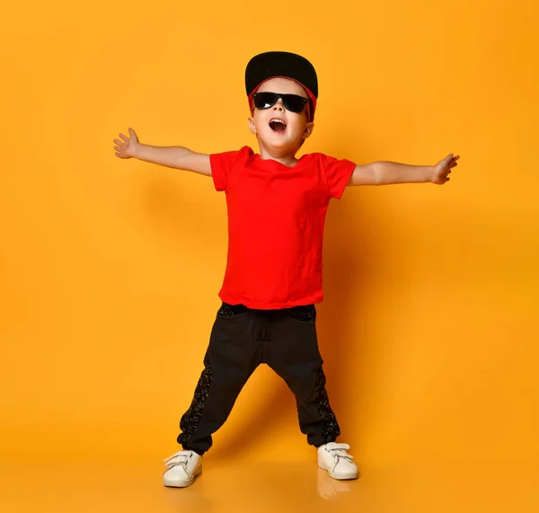 Młody chłopak w czerwonej koszulce i ciemnych spodniach, białych trampkach i śmiesznej czapce, pozujący na wolnej przestrzeni na żółtym tle. — Zdjęcie stockowe