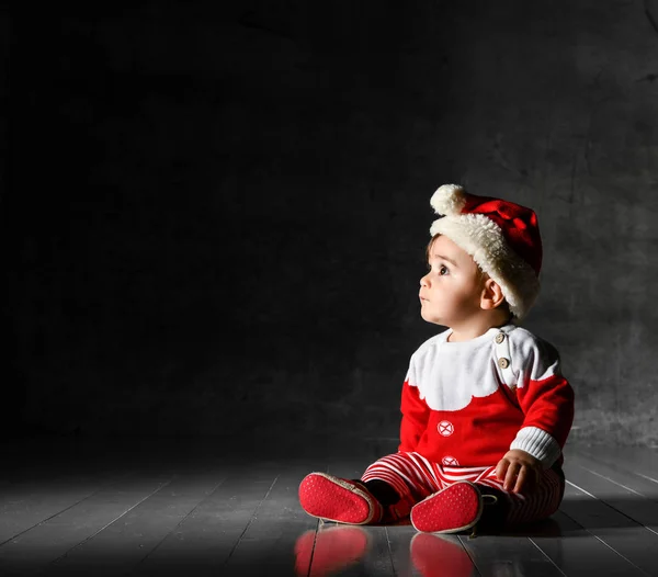 Маленький симпатичный маленький мальчик в красном свитере и кепке Санты, сидящий на темном фоне. — стоковое фото