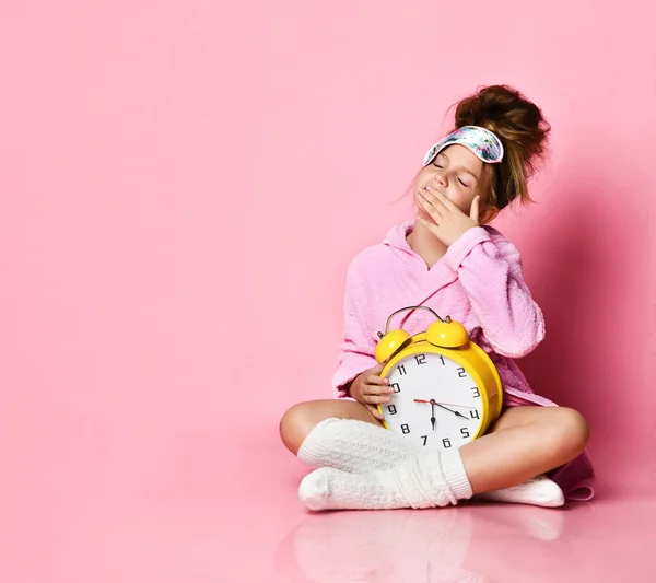 Yawning teenage girl holding an alarm clock while sitting in a housecoat on the floor.