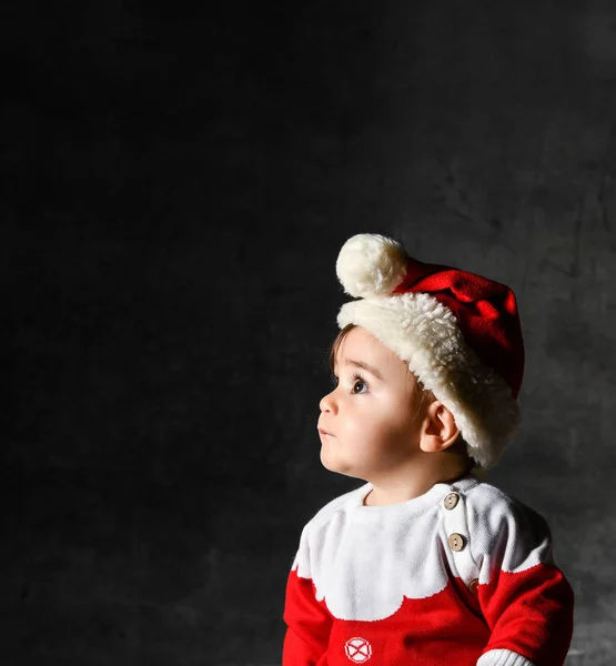Pequeno menino bonito em uma camisola vermelha e boné de Papai Noel, sentado em um fundo escuro. — Fotografia de Stock