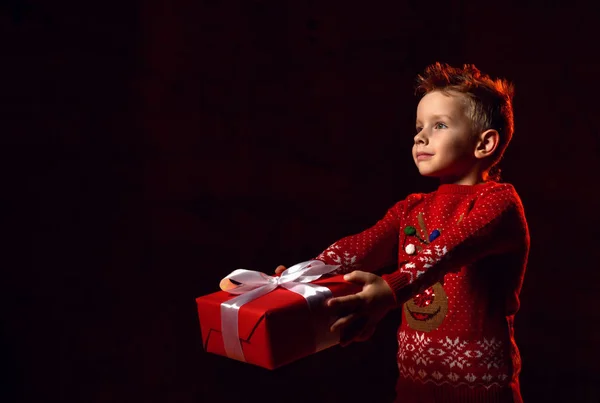 Petit garçon heureux dans un pull rouge avec une image d'un cerf de rudolphe tenant un cadeau du Père Noël — Photo