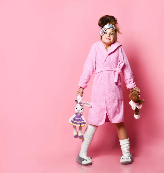 Grappig meisje in roze huisjurk en pluche sneakers houdt twee speelgoed — Stockfoto