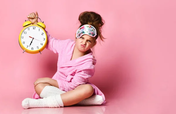 Yawning teenage girl holding an alarm clock while sitting in a housecoat on the floor. — Stock Photo, Image