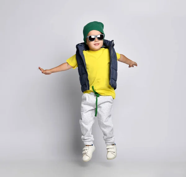 Elegante niño alegre 4 años de edad en pantalones de chándal, un sombrero verde, gafas y un chaleco abajo se divierte —  Fotos de Stock