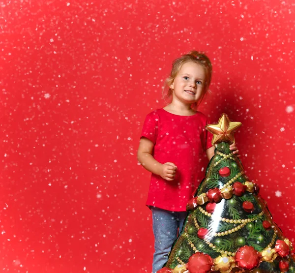 Menina criança feliz em uma camiseta vermelha, jeans azul segurando um balão de árvore de Natal em um fundo vermelho com espaço livre cópia . — Fotografia de Stock