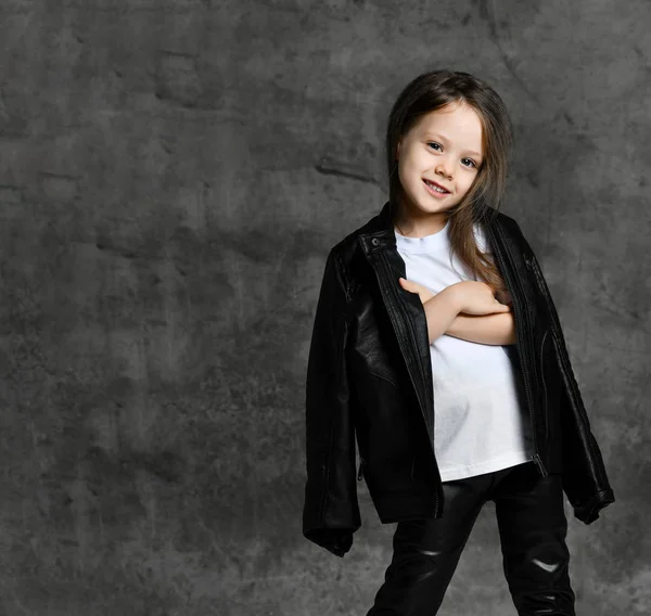Klein lachend schattig meisje in zwart-wit rock ster stijl casual kleding en witte sneakers staan over grijs beton achtergrond — Stockfoto