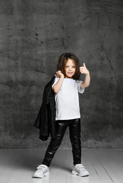 穿着黑白相间的休闲装和白色运动鞋的积极向上的小女孩站在那里，摆出大拇指姿势 — 图库照片