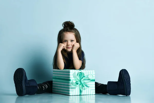 Klein lachend meisje in t-shirt, leren legging en donkerblauwe ugg laarzen zittend op de vloer met grote geschenkdoos — Stockfoto