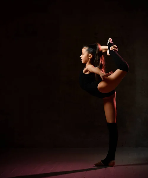 Bambina in una ginnastica nera ginnasta body esegue un esercizio. Equilibrio su una gamba con una presa . — Foto Stock