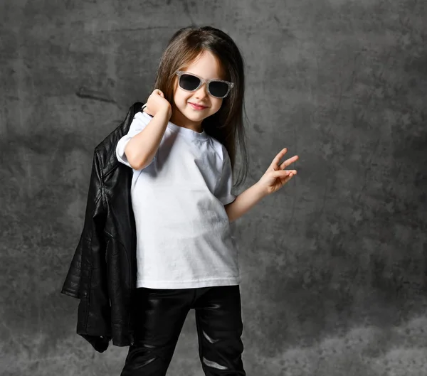 Klein positief meisje in zwart-wit rock stijl casual kleding en zonnebril staan en gelukkig voelen met opgeheven hand — Stockfoto