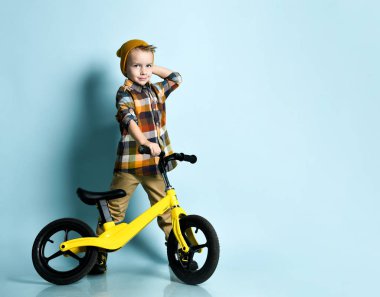 Parlak şık giyinen, spor ayakkabılı ve şapkalı pozitif çocuk mavi arka planda sarı bisikletin üzerinde oturuyor.