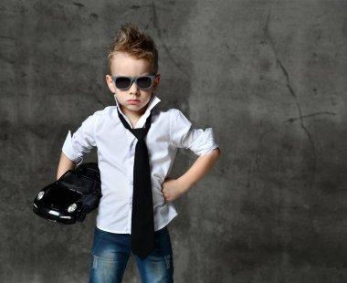 Havalı beyaz gömlekli, kot pantolonlu, kravatlı ve güneş gözlüklü gülümseyen çocuk elinde siyah bir oyuncak araba var.