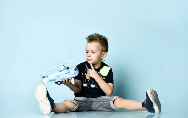 Usmívající se blondýnka chlapec ve stylovém ležérní oblečení sedí na podlaze s nohama rozházené a drží hračka vrtulník přítomen — Stock fotografie