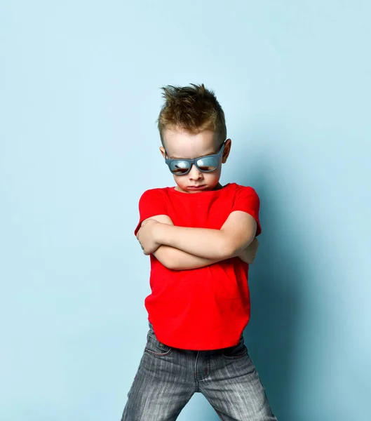 Серйозний хлопчик у яскравому стильному повсякденному одязі та сонцезахисних окулярах, що стоять над синім тлом — стокове фото