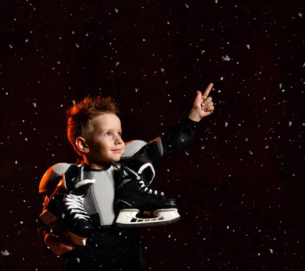 Seriózní chlapec v ochranné hokejové uniformě s bruslemi na krku stojící a natahující se přes tmavé pozadí se sněhové vločky — Stock fotografie