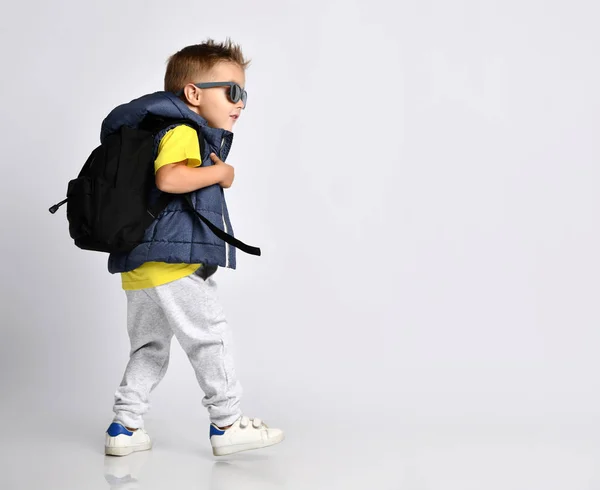 Atraktivní malý chlapec ve stylovém teplém oblečení s batohem na ramenou, baví na lehkém pozadí studia. — Stock fotografie