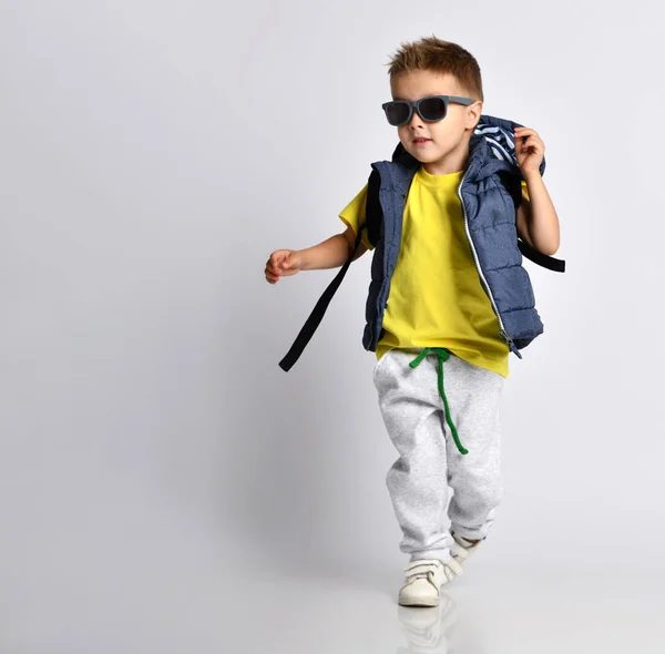 Attractive little boy in stylish warm clothes with a backpack on his shoulders, having fun on a light studio background. — Stock Photo, Image