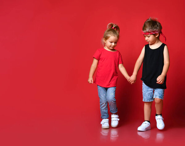 Small happy cute children boy and girl in stylish casual clothing go holding hands as couple over red background