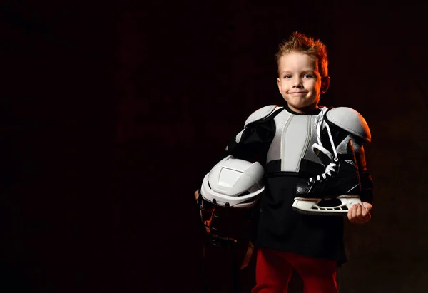 Menino loiro feliz em uniforme de hóquei com patins de hóquei e capacete de pé e sorrindo sobre fundo escuro — Fotografia de Stock