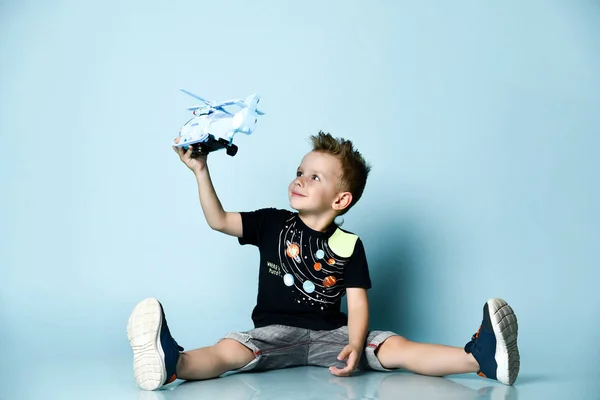 Lächelnder blonder Junge in stylischer Freizeitkleidung, der mit ausgebreiteten Beinen auf dem Boden sitzt und einen Spielzeughubschrauber in der Hand hält — Stockfoto