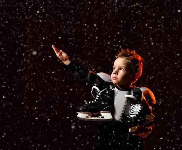Niño serio en uniforme de hockey protector con patines en el cuello de pie y alcanzando sobre fondo oscuro con copos de nieve — Foto de Stock