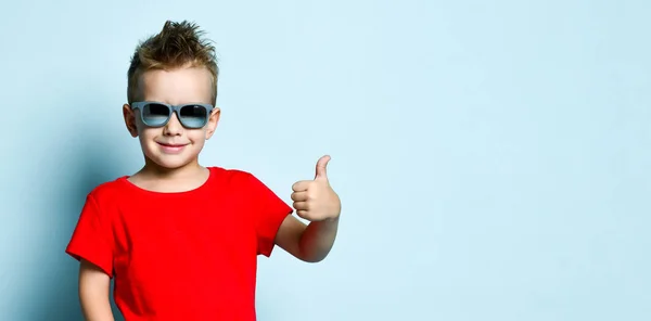 Rapaz sério em roupas casuais elegantes brilhantes e óculos de sol em pé sobre fundo azul — Fotografia de Stock