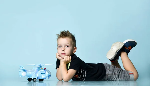 Positive blond boy in stylish casual clothing sitting on floor and holding toy helicopter present in hand over blue background