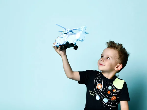 Pozytywne blond chłopiec w stylowej odzieży casual siedzi na podłodze i trzymając helikopter zabawka obecny w ręku nad niebieskim tle — Zdjęcie stockowe