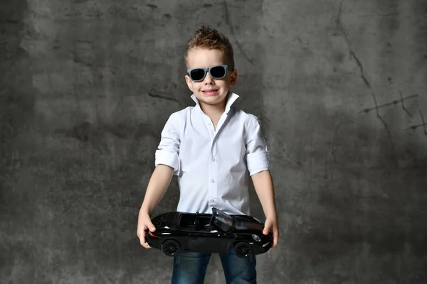 Garoto sorridente em elegante camisa branca casual, jeans, gravata e óculos de sol de pé e segurando carro de brinquedo preto presente na mão — Fotografia de Stock