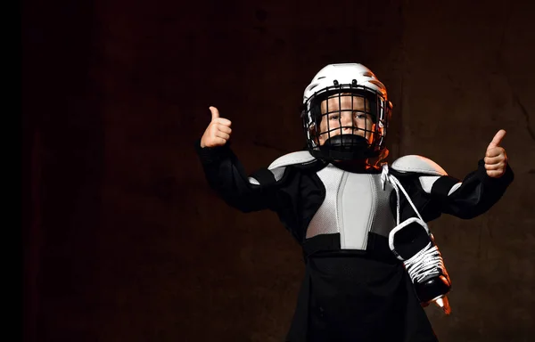 Pozytywny chłopiec w mundurze hokejowym i kasku ochronnym z łyżwami hokejowymi stojącymi i pokazującymi dobry znak kciuka — Zdjęcie stockowe