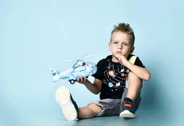 Pozitif sarışın çocuk şık günlük giysiler içinde yerde oturuyor ve elinde oyuncak helikopterle mavi arka planda bekliyor. — Stok fotoğraf