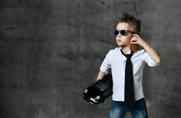 Vážný chlapec ve stylové bílé košili, džínách, kravatě a slunečních brýlích stojí, drží v ruce černé autíčko a volá na telefon — Stock fotografie