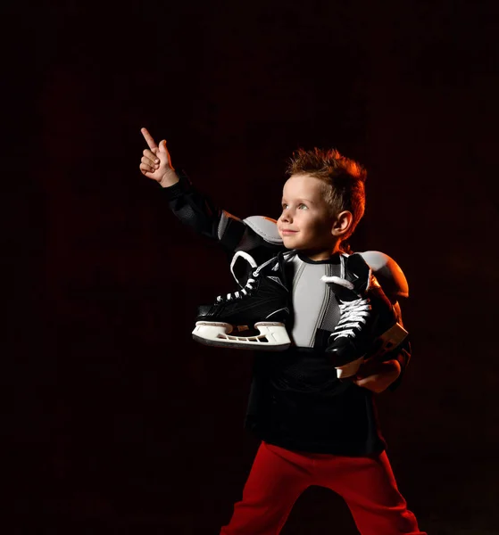 Blond jongetje in hockey uniform met hockey schaatsen op de hals staande en wijzen boven met vinger over donkere achtergrond — Stockfoto