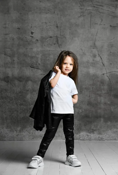 Klein positief meisje in zwart-wit rock stijl kleding en witte sneakers staan en poseren over grijs beton achtergrond — Stockfoto