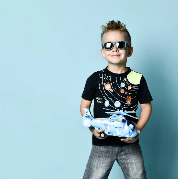 Menino loiro positivo em roupas casuais elegantes sentado no chão e segurando helicóptero de brinquedo presente na mão sobre fundo azul — Fotografia de Stock