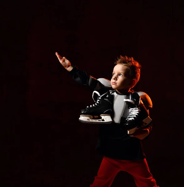 Menino loiro em uniforme de hóquei com patins de hóquei no pescoço em pé e apontando acima sobre fundo escuro — Fotografia de Stock
