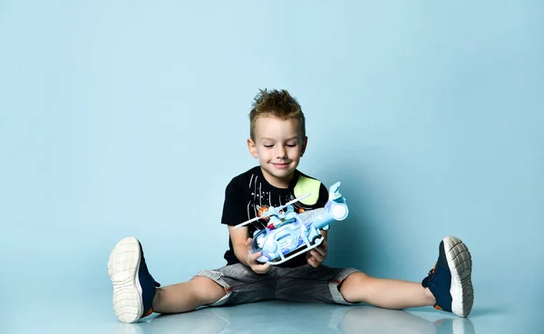 Usmívající se blondýnka chlapec ve stylovém ležérní oblečení sedí na podlaze s nohama rozházené a drží hračka vrtulník přítomen — Stock fotografie