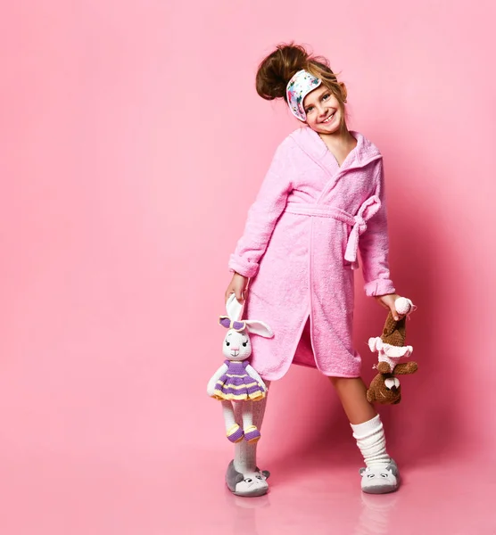 Grappig meisje in roze huisjurk en pluche sneakers houdt twee speelgoed — Stockfoto