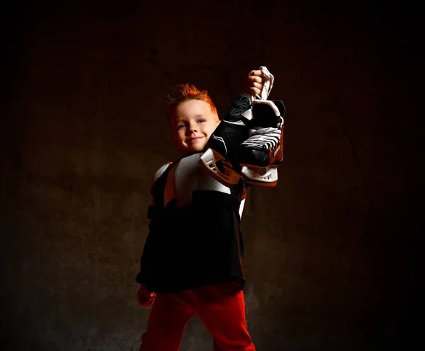 Blonďatý chlapec v hokejové uniformě s hokejové brusle na krku stojící a při pohledu na kameru přes tmavé pozadí — Stock fotografie