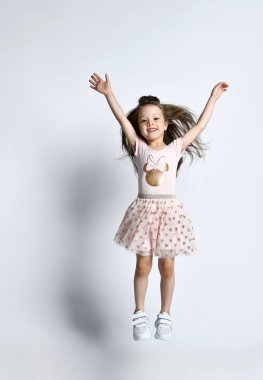 Small smiling cute girl in casual summer dress and sneakers jumping over white wall background