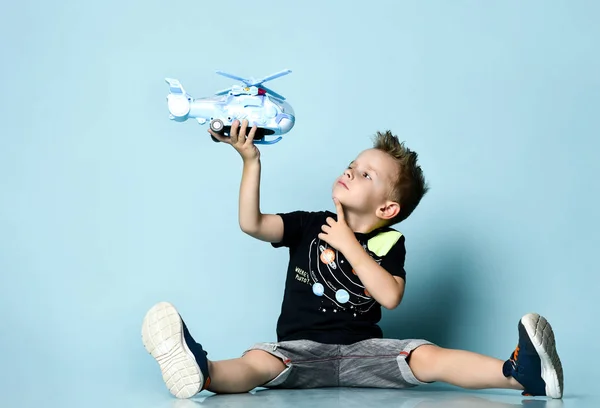 Blond chlapec ve stylovém ležérní oblečení sedí na podlaze a hrát si s hračkou vrtulník dárek přes modré pozadí — Stock fotografie