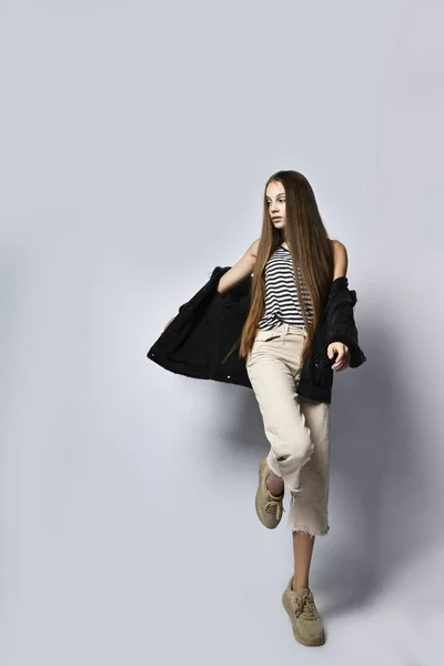 Подросток в полосатой футболке, черной джинсовой куртке, брюках и кроссовках. Она позирует изолировано на белом фоне. Закрыть — стоковое фото