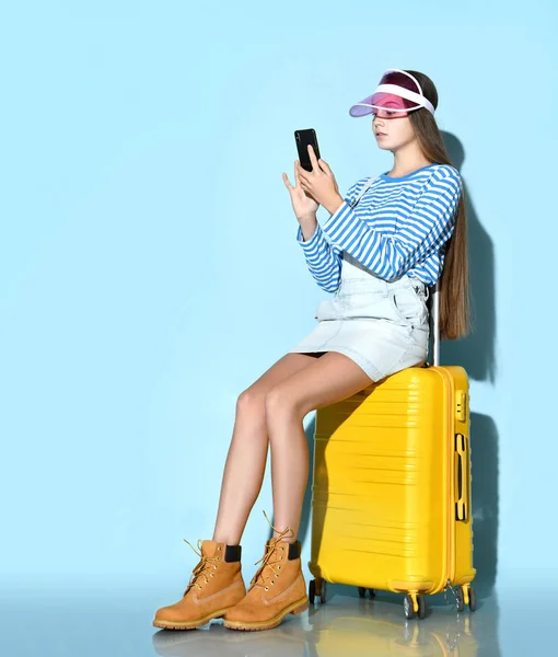 Teenage female in overall, sweatshirt, boots, sun visor cap. She using smartphone, sitting on yellow suitcase, blue background. — ストック写真