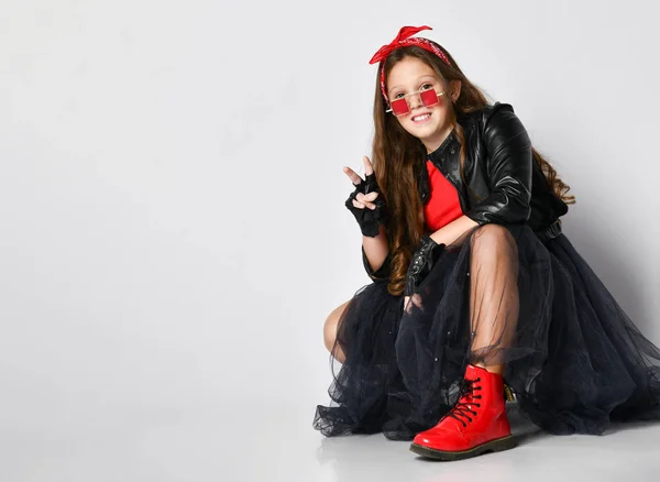 Jong lachend meisje in heldere rock stijl kleding, rode laarzen en accessoires zitten op de vloer met en wijzen met de vingers — Stockfoto