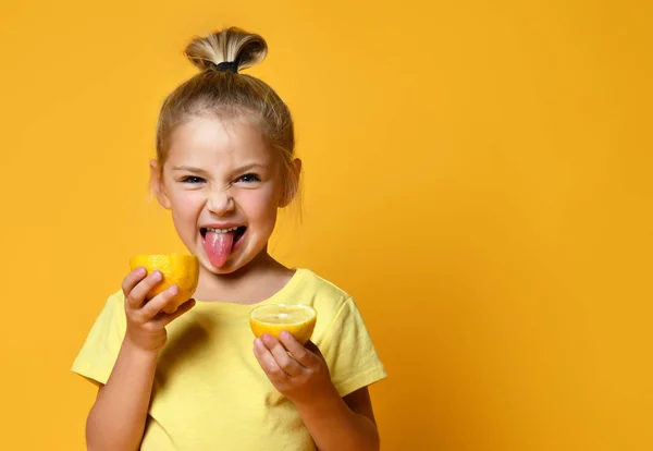 Klein lachend schattig meisje in geel t-shirt met helften van verse zure citroen fruit en tongzoenen over gele achtergrond — Stockfoto