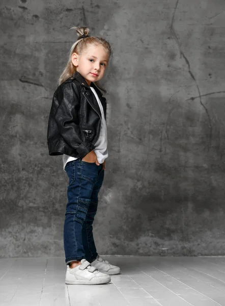 Klein lachend blond meisje in stijlvolle rock stijl zwart leren jas, blauwe jeans en witte sneakers staan met de handen in zakken — Stockfoto