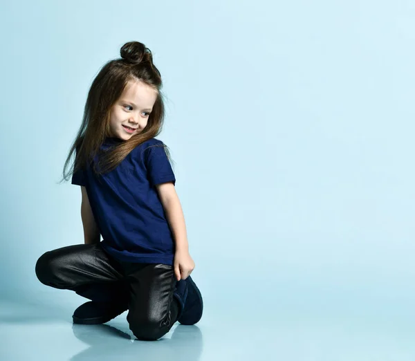 Klein lachend meisje met lang haar in casual t-shirt, leren broek en donkerblauwe ugg laarzen zittend op de vloer en naar beneden kijkend — Stockfoto