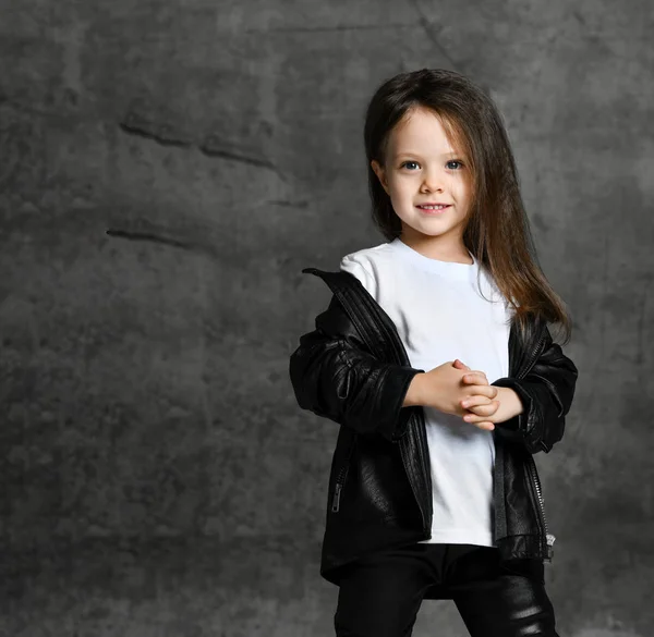Klein lachend schattig meisje in zwart-wit rock ster stijl casual kleding staan en kijken naar de camera over grijs beton achtergrond — Stockfoto